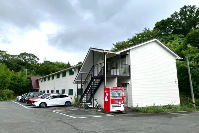 駐車場に面した白い2階建ての従業員寮の外観。建物の前には赤い自動販売機が置かれています。