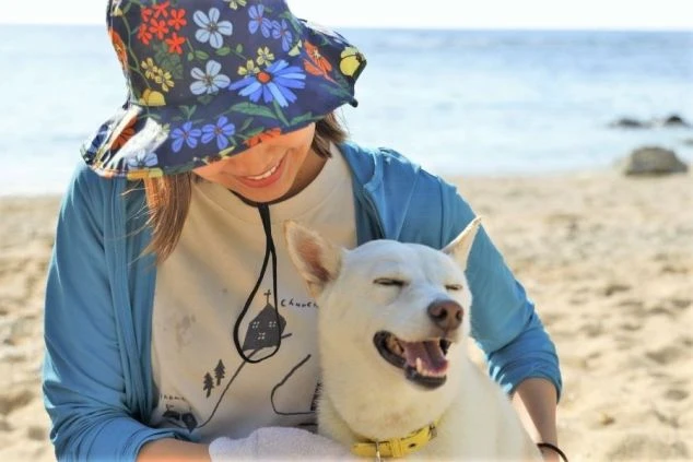 浜辺で、花柄の帽子をかぶった女性が白い犬を抱いて微笑んでいる様子。犬は目を細めて口を開け、笑っているように見える。背景は穏やかな海。