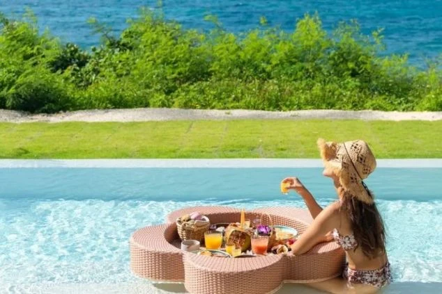 プールサイドで、麦わら帽子をかぶった女性が、水面に浮かんだバスケットに乗った朝食（パンケーキ、フルーツ、ジュースなど）を楽しんでいる。背景は青い海と緑の植栽。