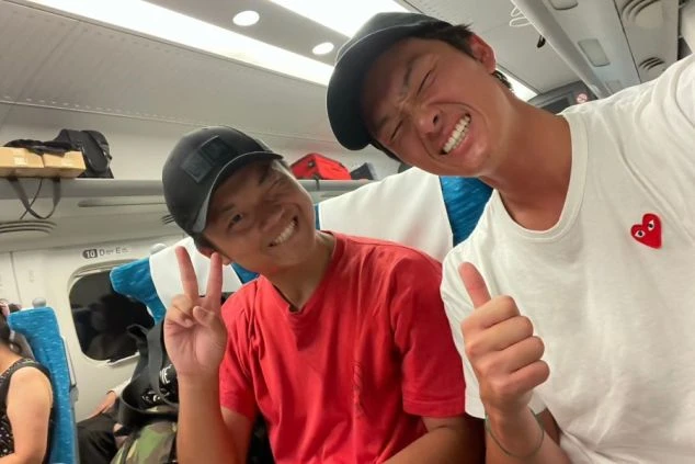 特急列車の車内で、リゾートバイト先の下田へ向かう笑顔の男性スタッフ2人。ピースサインやサムズアップで、これから始まる住み込みバイト生活への期待感があふれる自撮り写真。