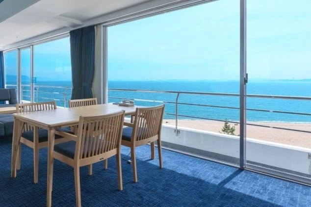 海に面したリゾートマンションまたはホテルの客室。全面ガラス窓から、青い海と砂浜が一望でき、室内には青いカーペット、木製のテーブルと椅子が置かれている。