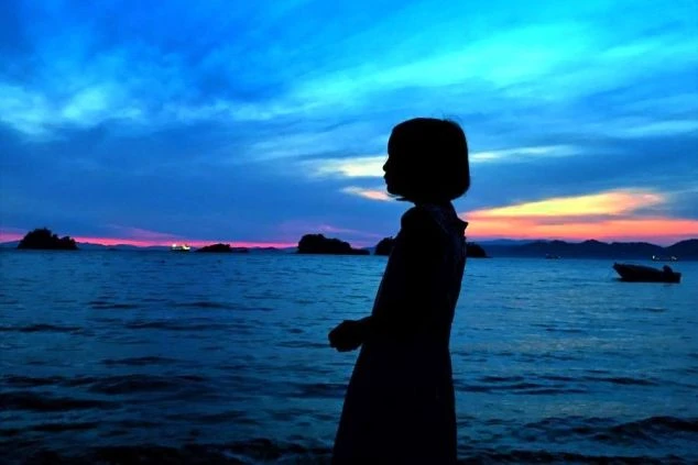 夕暮れの海辺に立つ人物のシルエット。空は濃い青色とオレンジ色に染まり、水面は暗い色調で波打っている。遠景にはいくつかの島影が見える。