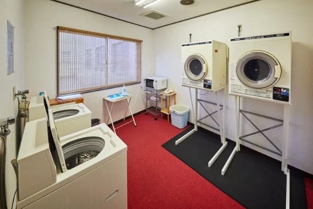 複数の洗濯機と乾燥機が並んだ、コインランドリーの画像。奥には電子レンジや、洗濯物を畳むための台が置かれている。リゾートバイトの寮の共用スペースにある、洗濯機の画像の画像。