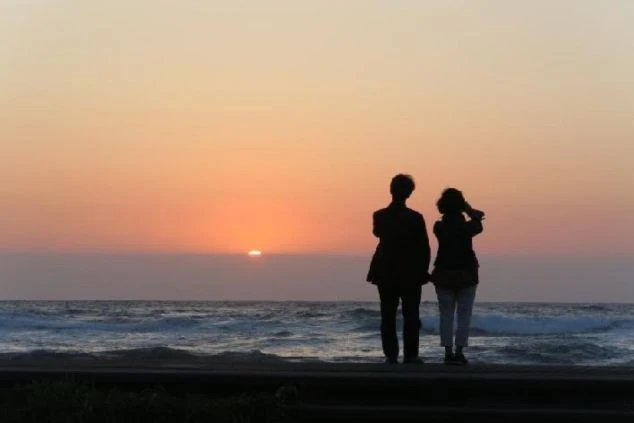 夕暮れの海辺で、2人のシルエットが夕日を眺めている画像。空と海がオレンジ色に染まり、波が打ち寄せる様子がわかる。リゾートバイトの休日に観光やデートを楽しんでいる風景の画像。