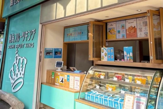 水色の外観が特徴的な「熱海フルーツキング」の店舗。ショーケースにはフルーツサンドやゼリーが並び、上部にはメニューが掲示されている。