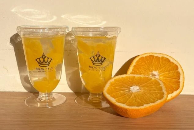 「熱海フルーツキング」のロゴが入ったグラスに入った、果肉たっぷりのフルーツゼリー。隣には半分にカットされたフレッシュなオレンジが2つ置かれている。