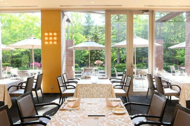 窓から緑豊かな庭園が見える、明るく開放的なレストラン。テーブルには食器がセッティングされ、リゾートバイトでホールスタッフや配膳の仕事に興味がある方に訴求する画像。