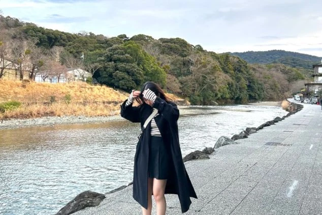 豊かな緑に囲まれた川辺で、コートを羽織り髪を整える女性の全身ショット。