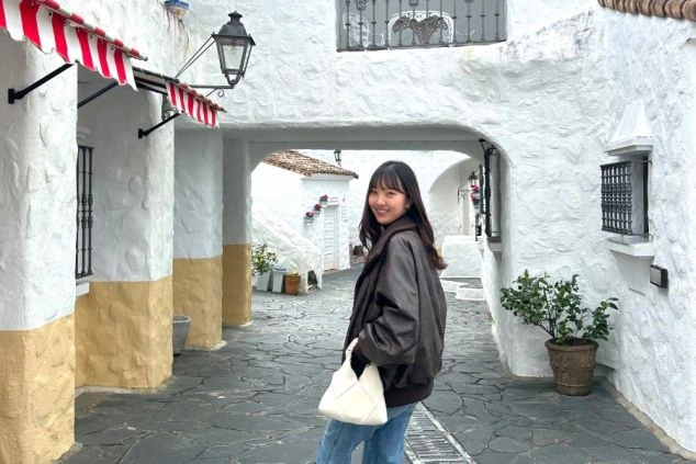 白壁の建物と石畳が続く異国情緒あふれる小道で、白いバッグを手に振り返り微笑む女性。