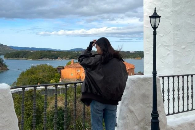 白壁のテラスから、穏やかな英虞湾とオレンジ色の屋根の建物を眺める女性の背中。