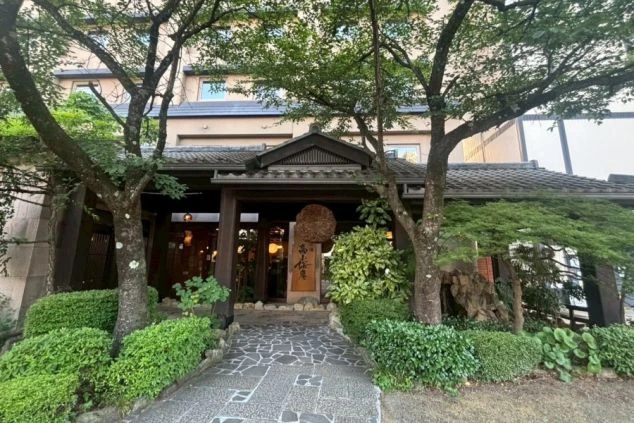 和風の玄関口と瓦屋根。両脇には大きな木と植栽があり、敷石が敷かれたアプローチの正面に「高山桜庵」の看板と大きな酒林が掛かっている。