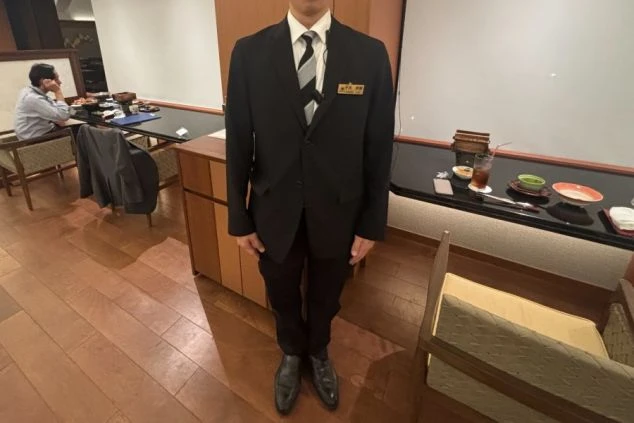 黒いスーツとストライプのネクタイを着用したホテルのレストラン・フロントスタッフの制服。落ち着いた雰囲気のレストランを背景に、リゾートバイトでホテルや旅館の接客業務に興味がある方に訴求する画像。