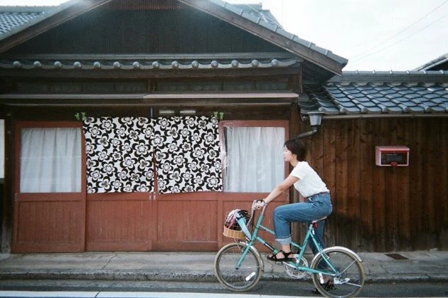 直島の風情ある古い日本家屋を背景に、若い女性が水色の自転車に乗って通りを移動している。