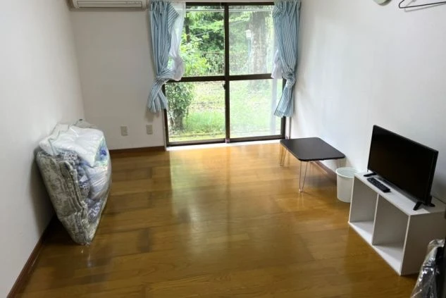 リゾートバイト寮の室内。フローリングの床に布団、テレビ、小さなテーブルが置かれており、窓からは豊かな緑が見える。