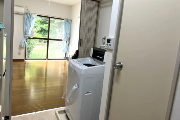 リゾートバイト寮にある洗濯機。玄関付近に設置されており、部屋の中にはフローリングの床と窓が見える。