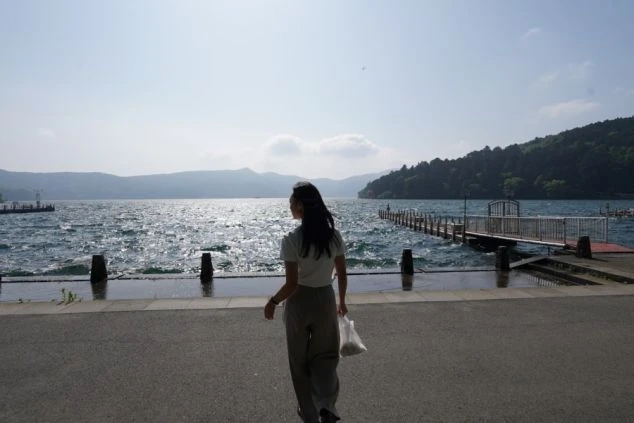 湖畔の道を歩く女性の後ろ姿。湖には波が立ち、奥には桟橋と緑豊かな山々が見える。
