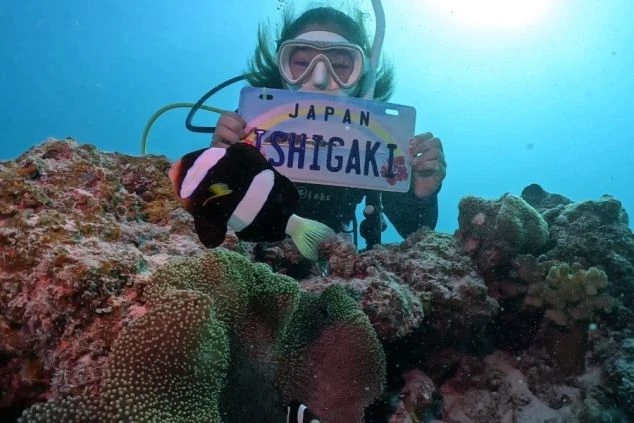 沖縄県石垣島の海中で、ダイビングスーツを着た人物が「JAPAN ISHIGAKI」と書かれたプレートを持ち、その手前にはクマノミが泳いでいる画像。石垣島の豊かな海中世界とアクティビティの楽しさ。