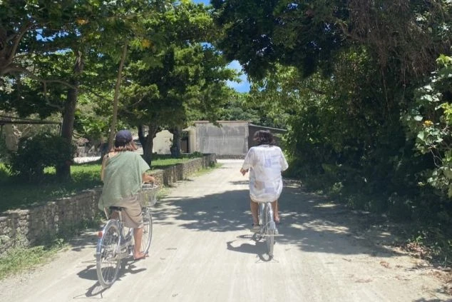 沖縄の竹富島で、木々の間を自転車で走る2人の人物の後ろ姿の画像。日差しが降り注ぎ、南国らしい開放的な雰囲気が感じられる。島のゆったりとした時間を象徴する風景。
