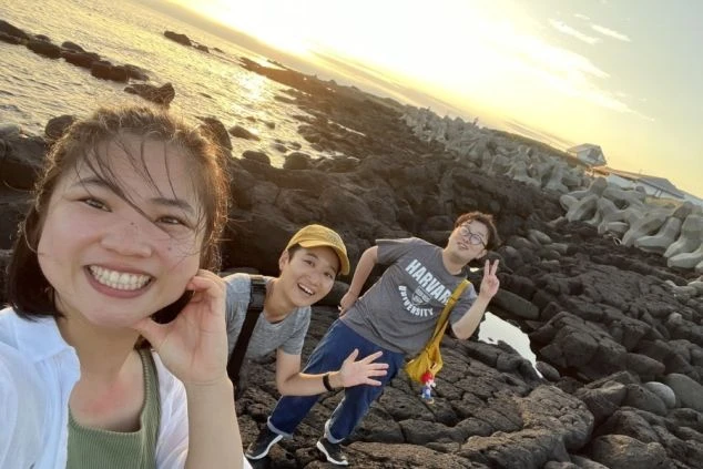 ワクトリでリゾートバイトしたスタッフが撮影した、北海道礼文島の夕焼けに染まる海岸で、笑顔でポーズをとる3人の若い男女の画像。岩場とテトラポッドが見える背景は、礼文島ならではの自然景観。