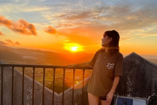 沖縄県石垣島の展望台から、夕焼け空を背景に笑顔でたたずむ女性の画像。地平線に沈む太陽が空と景色をオレンジ色に染め上げ、リゾートバイトで得られる感動的な体験や非日常感を表現している。