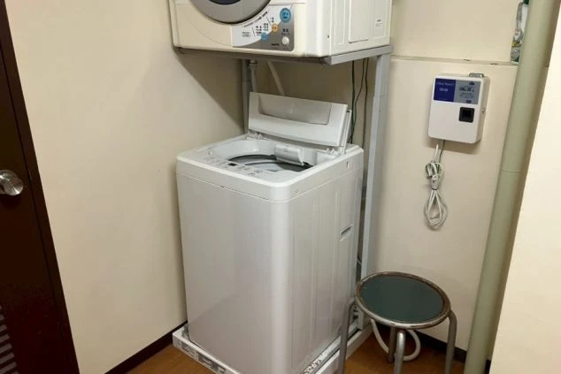 寮の洗濯スペース。全自動洗濯機と、その上に乾燥機が設置された、二段重ねの洗濯設備。右側にはタイマーまたは課金式の機器が見える。