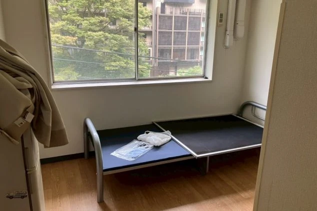 寮の居室。木のフローリングで、窓の外には緑と隣接する建物が見える。部屋には簡易なパイプ製のベッドフレームと、冷蔵庫らしき家電が置かれている。