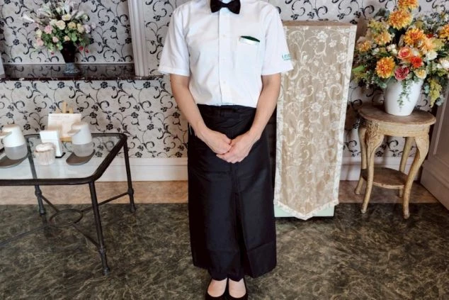 ホールの制服を着た女性。白いシャツに蝶ネクタイと黒いロングエプロンを着用している。