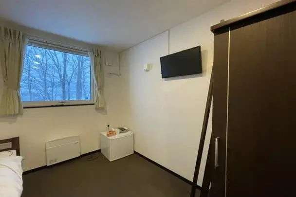 寮の居室の一角。壁には小型のテレビがかけられ、その横にはクローゼットと小型冷蔵庫がある。窓から差し込む外の光が強い。