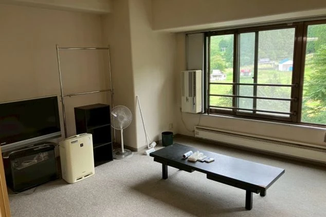 リゾートバイトの寮の室内。カーペット敷きの部屋に、テレビ、ハンガーラック、棚、扇風機が設置されている。窓からは外の景色が見え、快適な生活空間をアピール。
