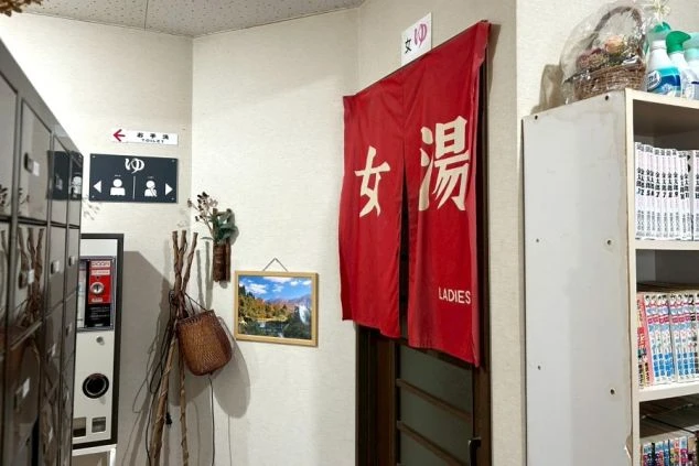 リゾートバイトの寮にある浴場入り口。「女湯」と書かれた赤い暖簾や、ロッカー、漫画本が並ぶ棚があり、共同浴場や娯楽スペースが充実していることをアピール。