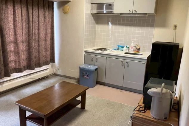 キッチンとリビングスペースが一体になっており、テーブルや調理器具、小型冷蔵庫が見える。