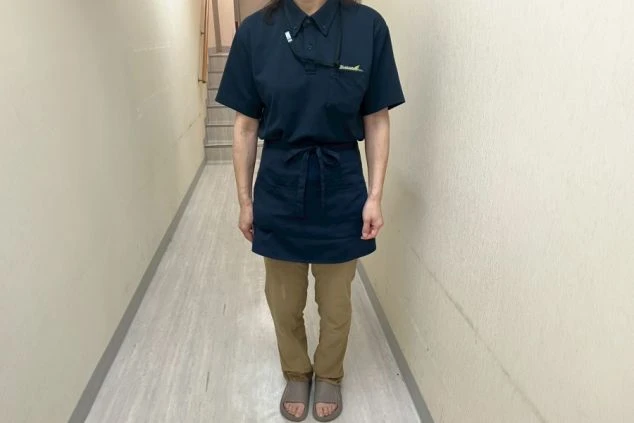 紺色のポロシャツとエプロン、ベージュのパンツを着用した女性スタッフの画像。首からはストラップ、腰からは鍵がぶら下がっている。リゾートバイトの仕事内容を表す、大浴場スタッフの制服の画像。