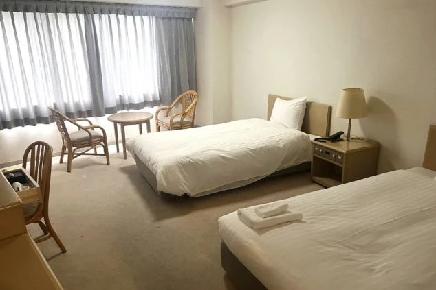 ワクトリの社員が出張で撮影した、リゾートバイトの客室寮の画像。ツインベッドが備え付けられた洋室で、デスク、椅子、サイドテーブル、ラタンチェアも配置されている。