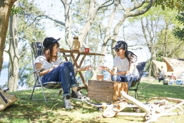 木々の下の芝生で、アウトドアチェアに座りワイングラスを交わす2人の女性の画像。奥にはテントも見え、キャンプやピクニックを楽しんでいる様子がうかがえる。