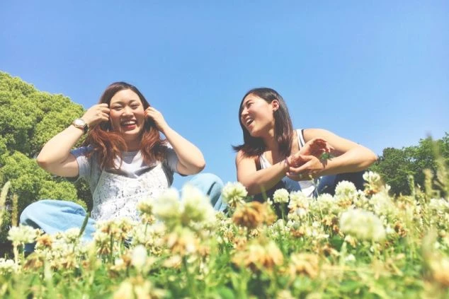 快晴の空の下、シロツメクサの咲く広場で笑顔で座り、楽しそうに話す2人の女性の画像。リゾートバイトの休日に、大自然の中でリラックスして過ごす様子を表現している。