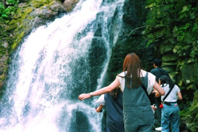滝壺近くで、勢いよく流れ落ちる滝を眺める人々の画像。特に女性が手を広げ、自然の雄大さを感じている様子。