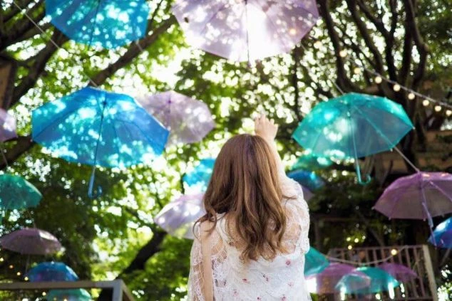 長野県軽井沢のハルニレテラスで、頭上に飾られた色とりどりの傘を見上げる女性の後ろ姿の画像。木漏れ日が差し込み、リゾートバイトの休日に訪れたい、幻想的で美しい観光スポットを表現している。