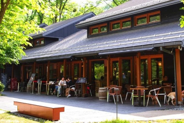 軽井沢のベーカリーの外観の画像。木目を基調とした落ち着いた雰囲気の建物で、テラス席では人々が食事を楽しんでいる。緑豊かな環境に溶け込むリゾート施設で、リゾートバイトの職場である。