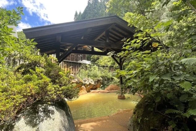 緑豊かな自然の中に設置された露天風呂の画像。浴槽は岩で囲まれており、屋根が付いている。リゾートバイトの寮やホテルの福利厚生としても利用できる、温泉の画像。