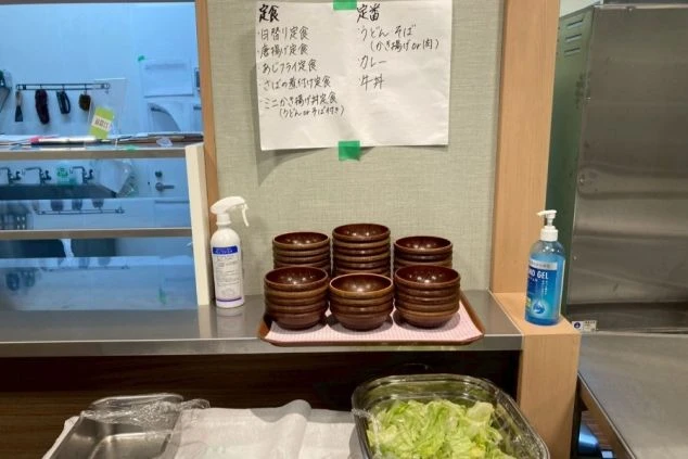 スタッフ食堂のカウンター。定食メニューが書かれた紙が貼られ、手前には重ねられた食器とサラダ、消毒液が置かれている。