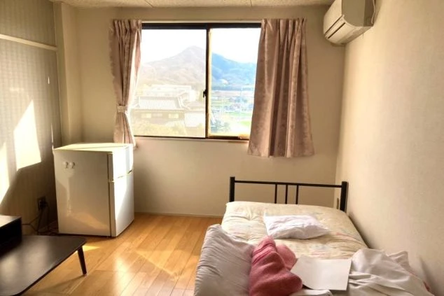 リゾートバイトの社員寮の部屋を表現するイメージ画像。窓から山々が見える、ベッドと冷蔵庫が備え付けられたシンプルな個室。