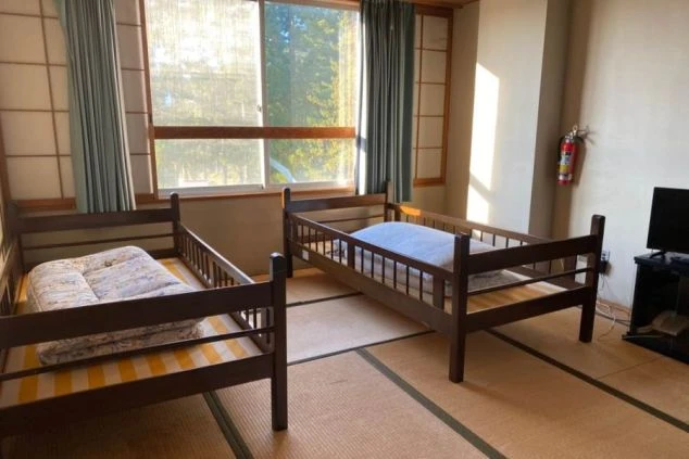 リゾートバイトの寮の相部屋。2つのベッドが並び、窓から光が差し込んでいる。和室の畳敷きの部屋で、テレビやヒーターも完備。