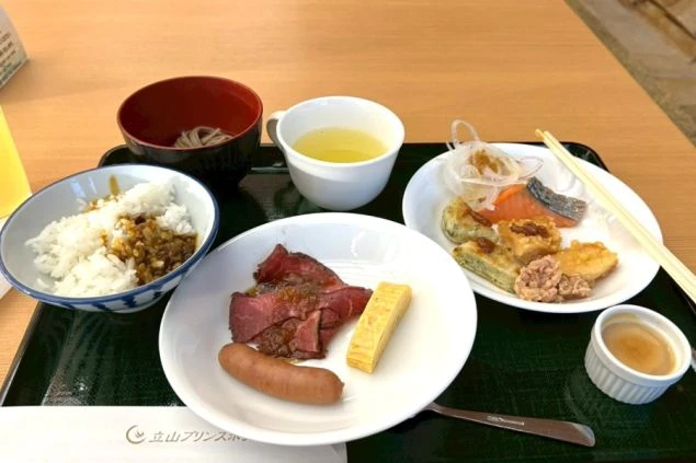 お盆にのった朝食。ローストビーフ、卵焼き、ソーセージ、焼き魚や揚げ物、ご飯、そば、スープが並ぶ。