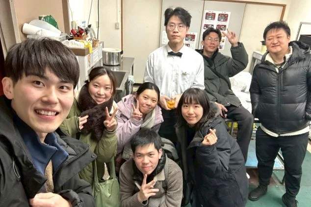 厨房のような室内で、冬服や制服を着た男女8人が笑顔で集まっている集合写真。ピースサインや指ハートを作っている。