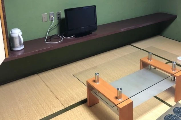 畳敷きの和室の隅に、壁に沿って設置された長い棚の上に小さなテレビと電気ケトルが置かれている画像。壁にはコンセントがあり、リゾートバイトの寮で過ごすプライベートな空間のイメージ。