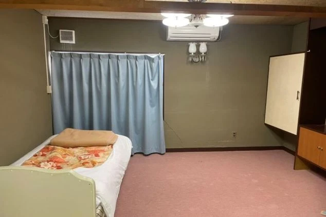 ピンク色のカーペットが敷かれた洋室に、ベッドが1台と壁にはエアコン、天井には照明が写っている画像。部屋の隅には収納家具も見え、リゾートバイトの個室寮で快適に過ごせる洋室の寝室のイメージ。