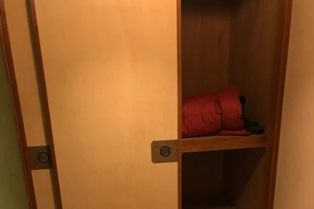 木製の引き戸タイプの収納が開いており、中に赤い寝袋のようなものが収められている画像。リゾートバイトの寮に備え付けられた、荷物や布団などを収納できる押し入れ(収納スペース)のイメージ。