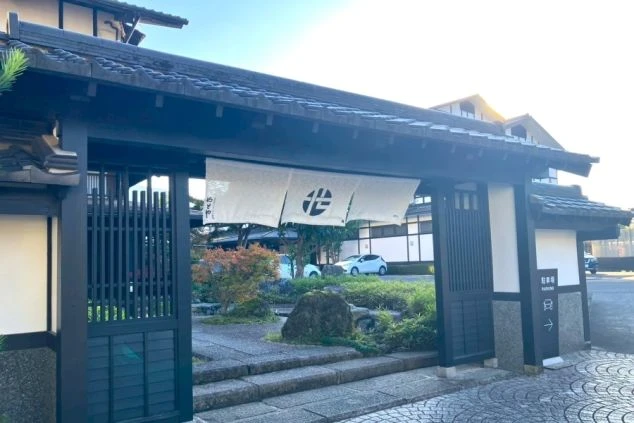 旅館の黒い門構え。石畳と植栽の先に建物が見え、門に旅館名が入った白い暖簾がかかっている。
