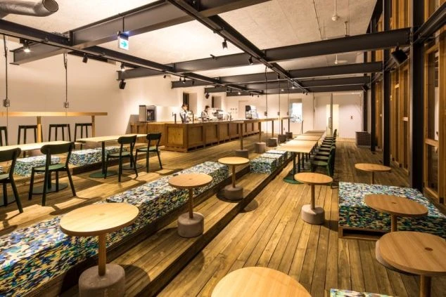 木目調の床とモダンなデザインのテーブルと椅子が並ぶ、広々としたカフェスペースの画像。奥にはカウンターがあり、ゆったりと過ごせるリゾート施設の共有空間のイメージ。