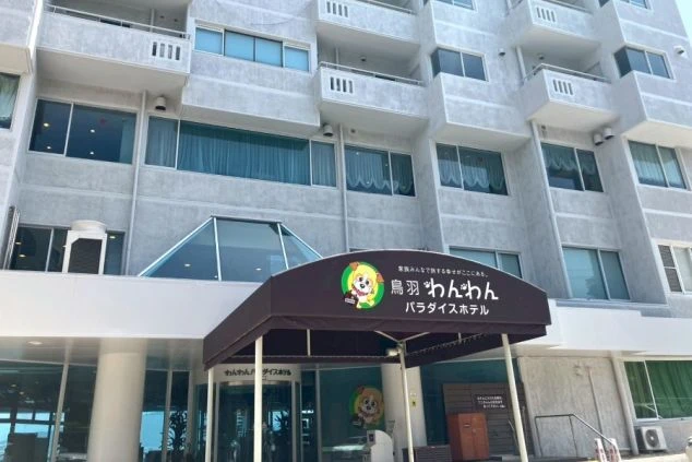 旅館またはホテル「鳥羽 わんわんパラダイスホテル」の外観。コンクリート造りの建物で、中央のエントランスには犬のマスコットキャラクターのロゴが入った庇（ひさし）が突き出ている。
